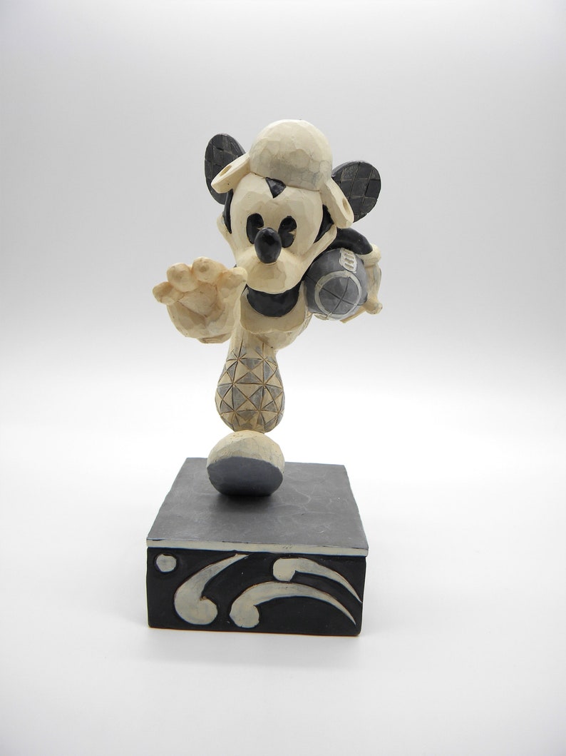 Puede incluir: Una figura tallada de Mickey Mouse con uniforme de b&eacute;isbol, sosteniendo una pelota de b&eacute;isbol. La figura es principalmente crema y negra, con una gorra de b&eacute;isbol y un dise&ntilde;o estampado en la pierna. Se encuentra sobre una base rectangular negra con detalles decorativos blancos.
