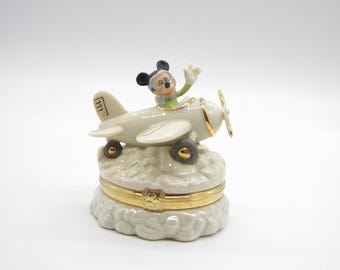 May include: A cream-colored ceramic music box featuring Mickey Mouse in a biplane. The plane has a gold propeller and sits atop a cloud-shaped base with a gold band. Mickey waves from the cockpit. The wheels are dark brown.