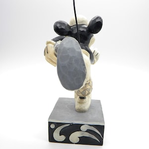 Puede incluir: Figura de madera de Mickey Mouse en una pose din&aacute;mica, con una pierna extendida. La figura es principalmente negra y crema, con una base gris y un acento negro. La figura tiene un elemento negro en forma de antena que se extiende hacia arriba.