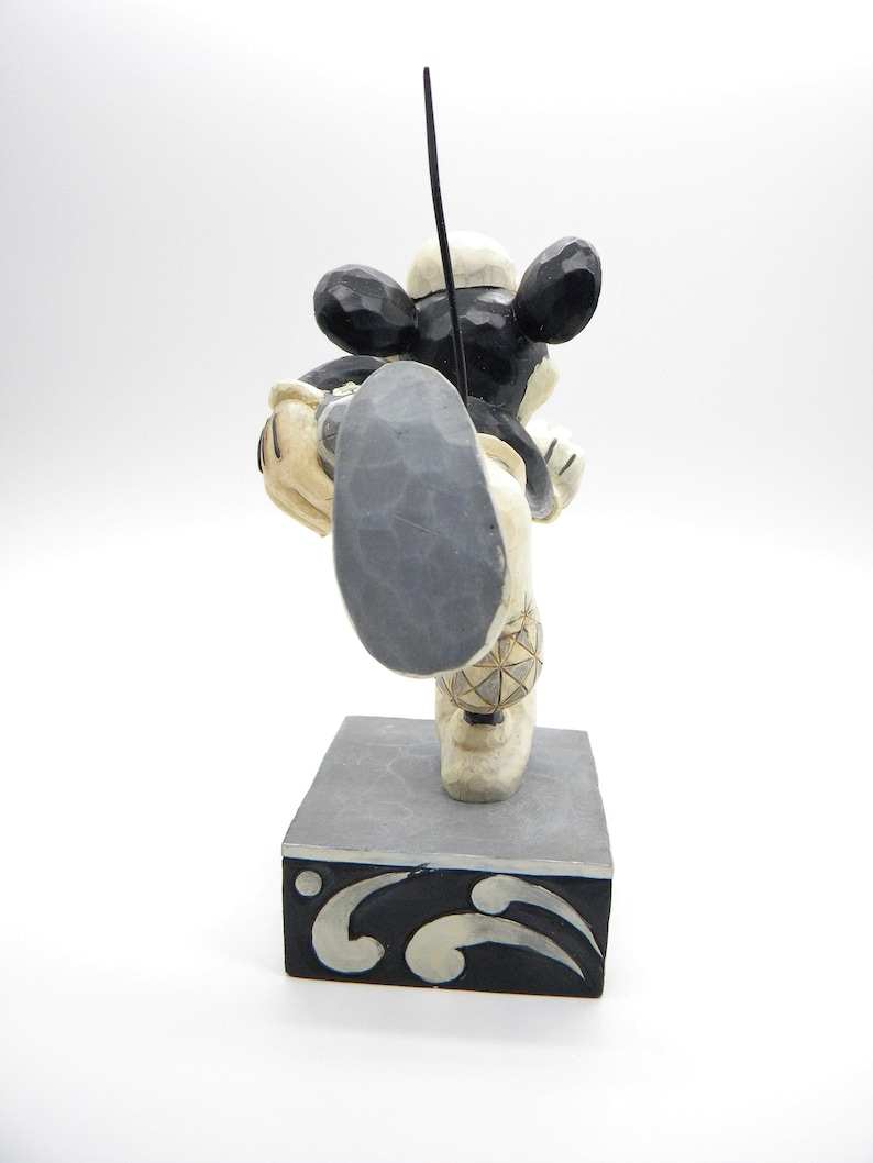 Puede incluir: Figura de madera de Mickey Mouse en una pose din&aacute;mica, con una pierna extendida. La figura es principalmente negra y crema, con una base gris y un acento negro. La figura tiene un elemento negro en forma de antena que se extiende hacia arriba.