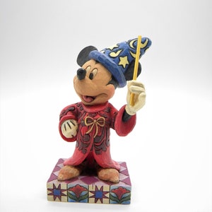 Op de afbeelding: Een beeldje van Mickey Mouse als tovenaar, met een blauwe hoed met gele sterren en een gele toverstaf. De rode mantel heeft bruine details en het beeldje staat op een patroonbasis. Een charmant verzamelobject.