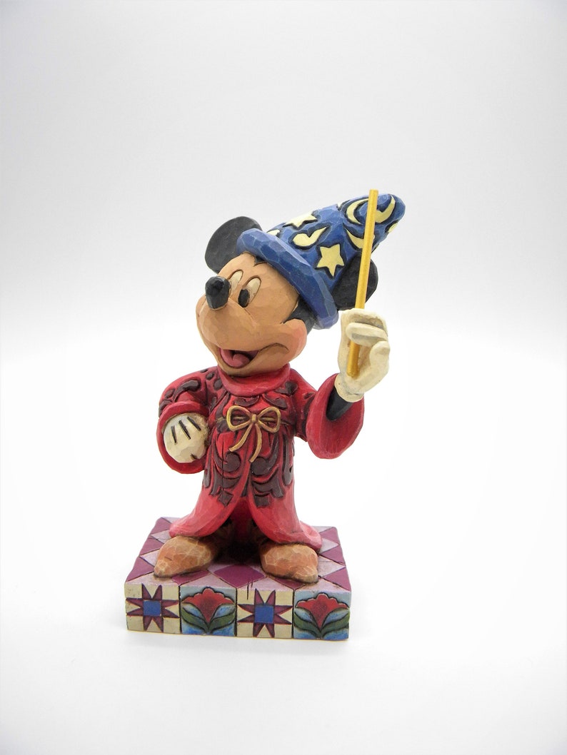 Op de afbeelding: Een beeldje van Mickey Mouse als tovenaar, met een blauwe hoed met gele sterren en een gele toverstaf. De rode mantel heeft bruine details en het beeldje staat op een patroonbasis. Een charmant verzamelobject.
