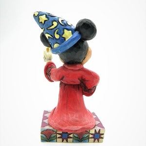 Op de afbeelding: Een beeldje van Mickey Mouse als tovenaar, met een blauwe hoed met gele sterren en manen. De figuur draagt een rode mantel en houdt een kaars vast. Het staat op een decoratieve basis met bloemen en geometrische patronen.
