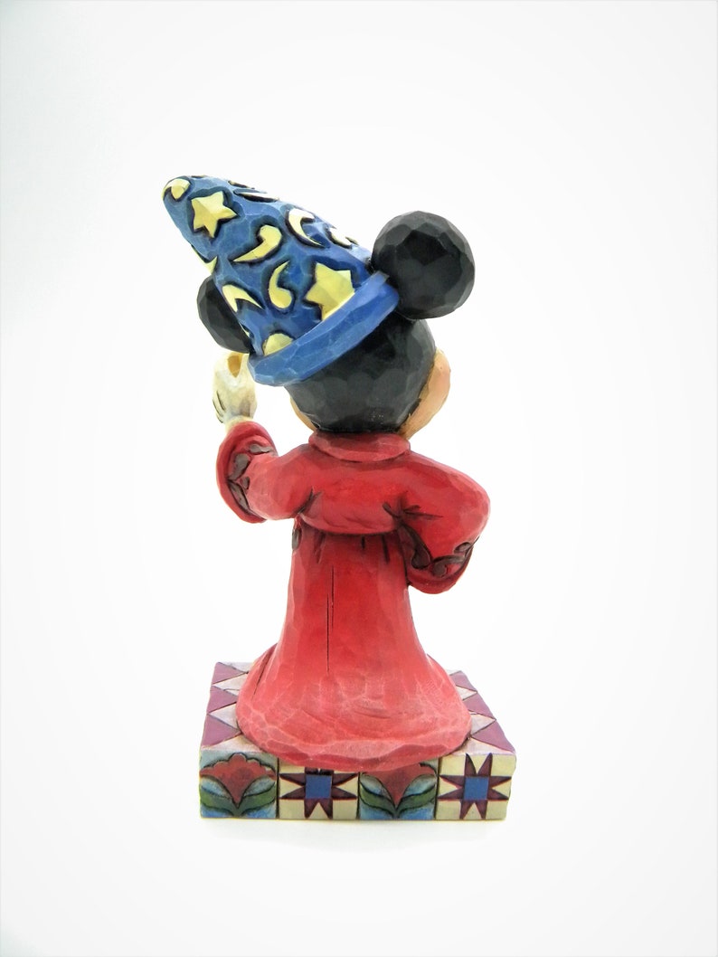 Op de afbeelding: Een beeldje van Mickey Mouse als tovenaar, met een blauwe hoed met gele sterren en manen. De figuur draagt een rode mantel en houdt een kaars vast. Het staat op een decoratieve basis met bloemen en geometrische patronen.