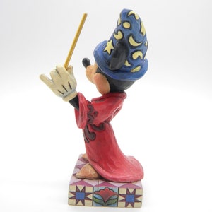 Op de afbeelding: Een gesneden beeldje van Mickey Mouse als tovenaar, met een blauwe hoed met gele sterren en een rode jurk. Mickey houdt een gele toverstaf vast en staat op een decoratieve basis met een bloemenmotief.