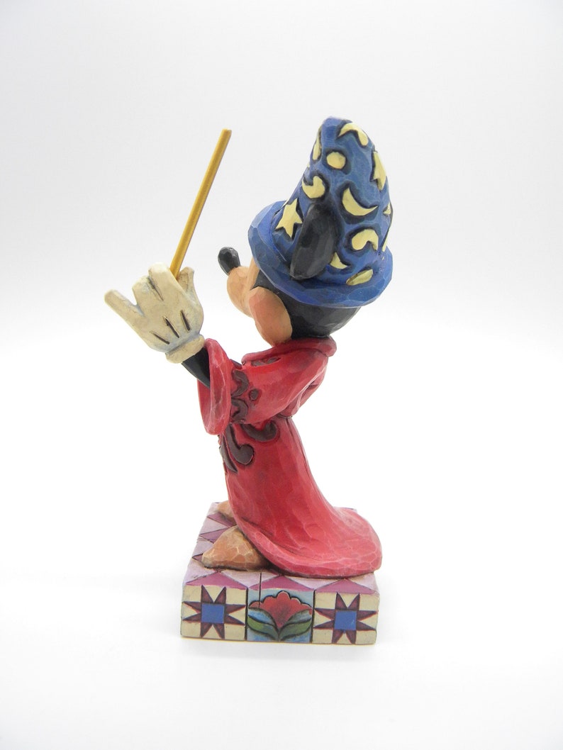 Op de afbeelding: Een gesneden beeldje van Mickey Mouse als tovenaar, met een blauwe hoed met gele sterren en een rode jurk. Mickey houdt een gele toverstaf vast en staat op een decoratieve basis met een bloemenmotief.