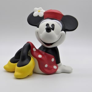 May include: A ceramic figurine of Minnie Mouse wearing a red dress with white polka dots, a red hat, and a white flower. She is sitting with her legs crossed and smiling.