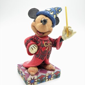 Op de afbeelding: Een Mickey Mouse-beeldje verkleed als tovenaar, met een blauwe hoed met sterren en een rode jurk. Het beeldje houdt een gouden toverstaf vast en staat op een patroonbasis. De figuur is ongeveer 13 cm hoog.