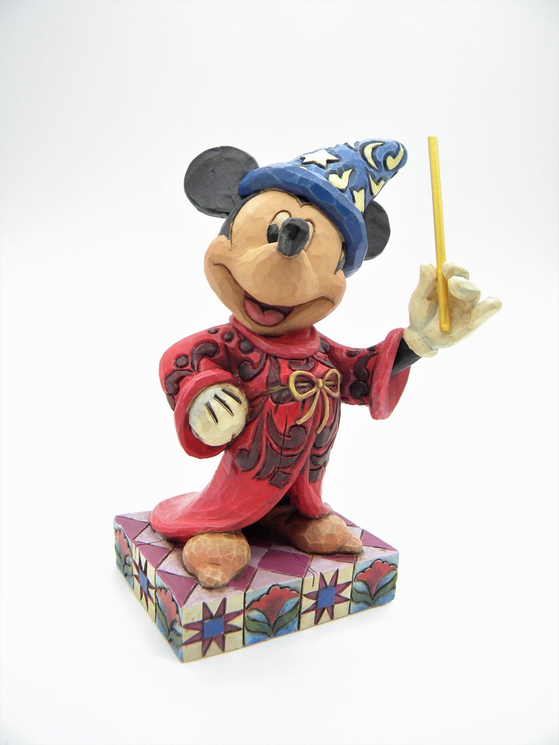 Op de afbeelding: Een Mickey Mouse-beeldje verkleed als tovenaar, met een blauwe hoed met sterren en een rode jurk. Het beeldje houdt een gouden toverstaf vast en staat op een patroonbasis. De figuur is ongeveer 13 cm hoog.