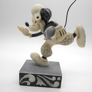 Puede incluir: Una figura tallada de Mickey Mouse con uniforme de f&uacute;tbol, en plena acci&oacute;n. La figura es principalmente de color crema con detalles en negro y un bal&oacute;n de f&uacute;tbol gris. Se encuentra sobre una base decorativa gris y negra.