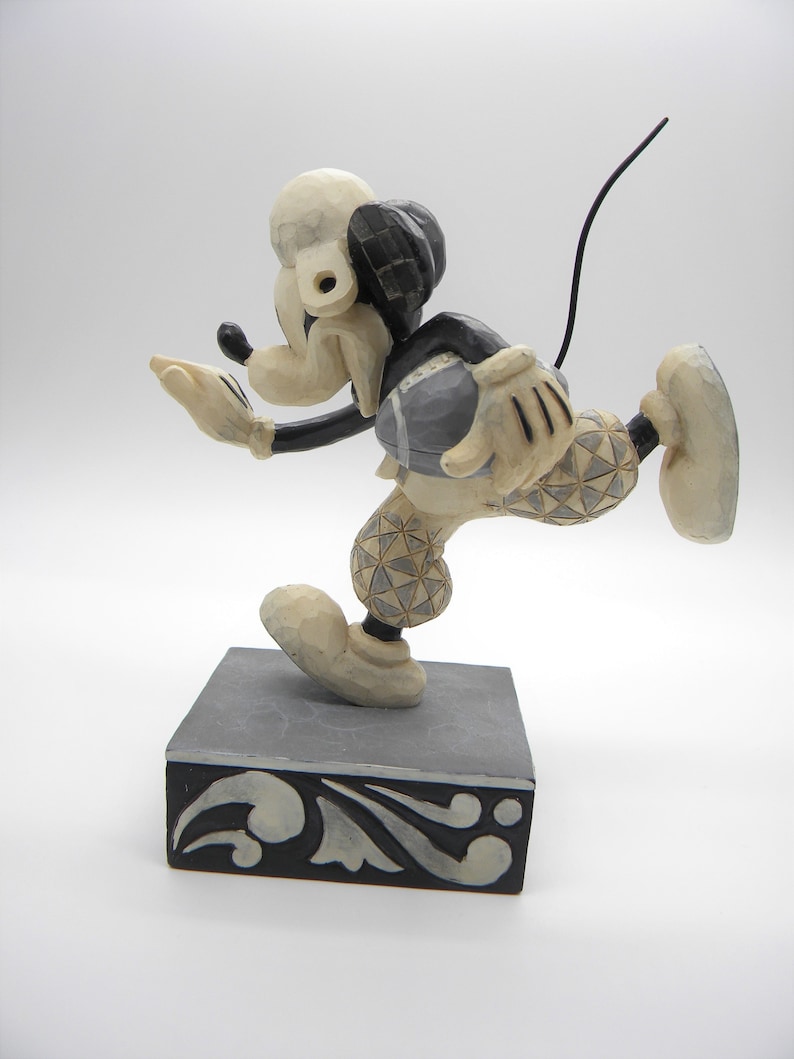 Puede incluir: Una figura tallada de Mickey Mouse con uniforme de f&uacute;tbol, en plena acci&oacute;n. La figura es principalmente de color crema con detalles en negro y un bal&oacute;n de f&uacute;tbol gris. Se encuentra sobre una base decorativa gris y negra.