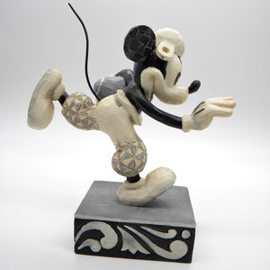 Puede incluir: Figura de Mickey Mouse en pose din&aacute;mica, con una pierna extendida y brazos extendidos. La figura es principalmente crema con detalles en negro, incluyendo las orejas y la cola. Se encuentra sobre una base decorativa gris y negra.