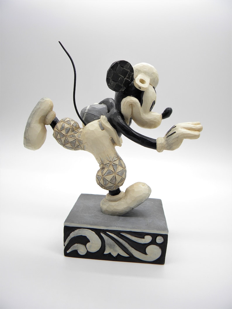 Puede incluir: Figura de Mickey Mouse en pose din&aacute;mica, con una pierna extendida y brazos extendidos. La figura es principalmente crema con detalles en negro, incluyendo las orejas y la cola. Se encuentra sobre una base decorativa gris y negra.
