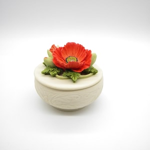 May include: A white ceramic trinket box with a floral design. The lid features a red poppy flower with green leaves.