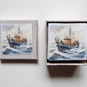 Könnte beinhalten: Zwei quadratische Schachteln mit nautischem Thema. Eine ist geschlossen, die andere offen und zeigt einen Stapel Karten. Jede Karte und der Deckel der geschlossenen Schachtel zeigen ein Aquarellgemälde eines Fischerbootes auf See in Blau, Orange und Weiß.