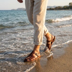 May include: Brown leather sandals with multiple straps and a buckle closure. The sandals are being worn while walking on a sandy beach with the ocean in the background. The person is wearing beige pants.