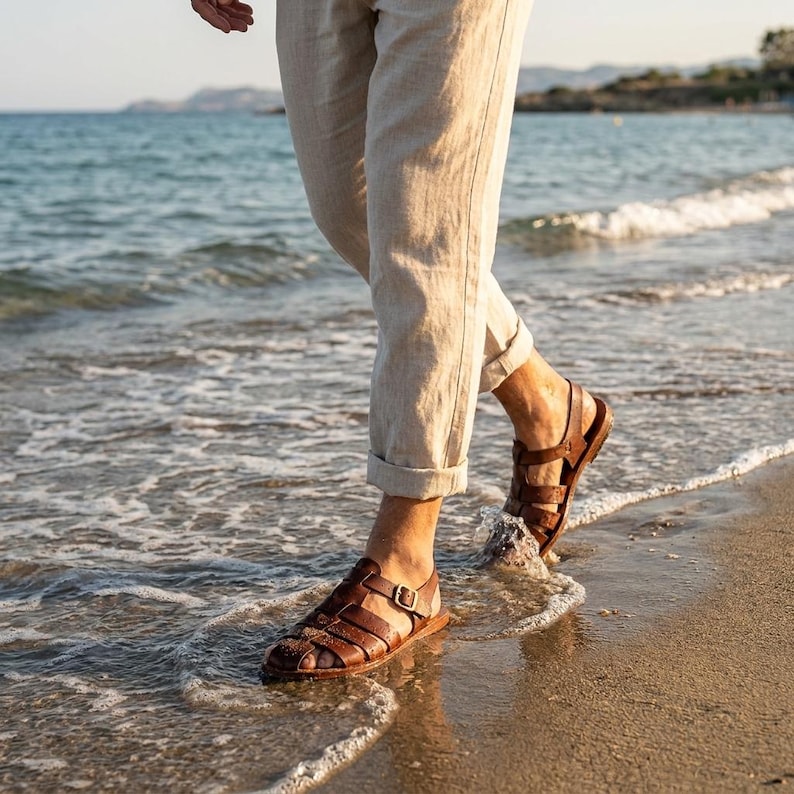 May include: Brown leather sandals with multiple straps and a buckle closure. The sandals are being worn while walking on a sandy beach with the ocean in the background. The person is wearing beige pants.