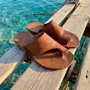 May include: A pair of brown leather sandals with a toe loop design. The sandals are placed on a wooden dock with the ocean in the background. The sandals appear to be handmade.