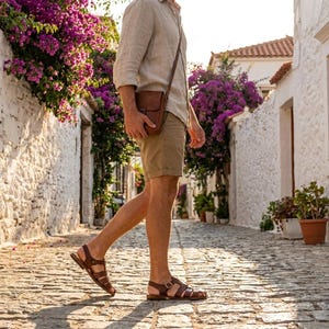 May include: A person walking down a cobblestone street wearing brown leather sandals, khaki shorts, and a light-colored button-down shirt. A brown leather crossbody bag is worn over the shoulder. The background features white buildings and purple flowers.