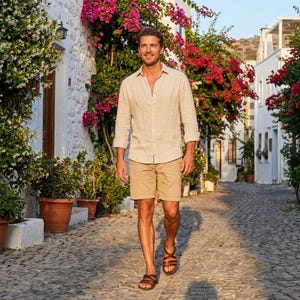 May include: A man wearing a beige linen shirt, khaki shorts, and brown leather sandals walks down a cobblestone street. The scene is framed by white buildings and vibrant pink flowers, creating a summery, relaxed vibe.