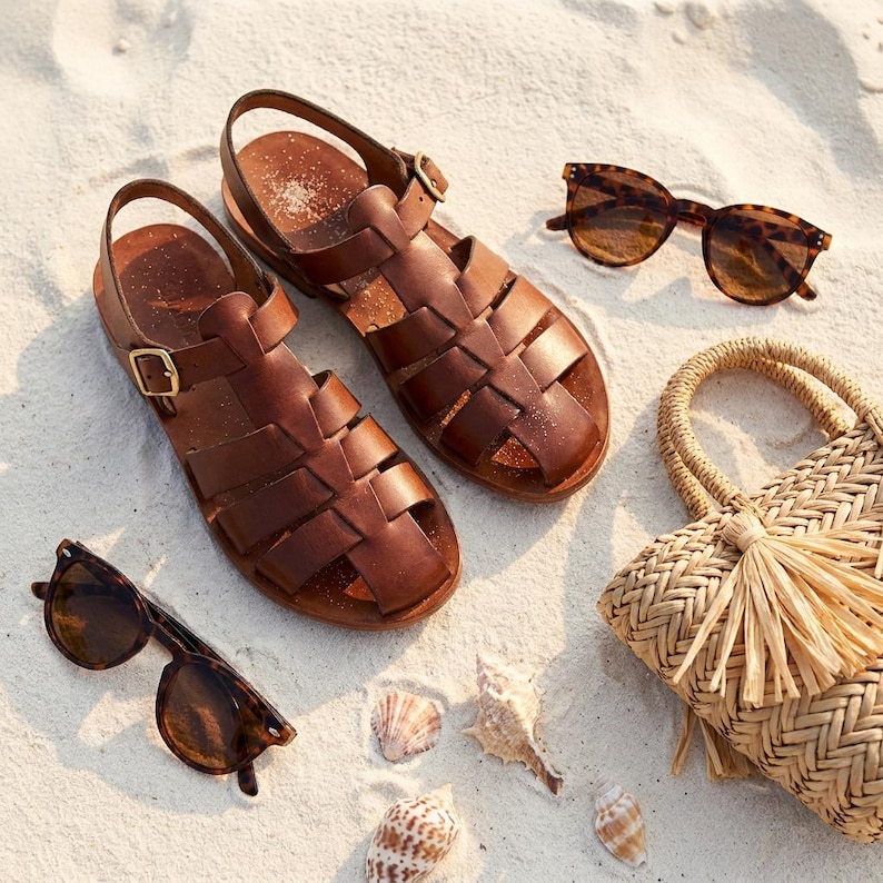 May include: A pair of brown leather sandals with a woven design, gold buckles, and a strap. Two pairs of tortoiseshell sunglasses, a woven straw handbag with a tassel, and seashells are arranged on a sandy surface.
