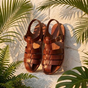 May include: A pair of brown leather sandals with a cage-like design. The sandals have a strap around the ankle and a buckle closure. The shoes are displayed against a white stone wall with green foliage.