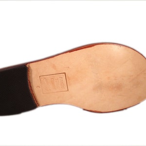 May include: Bottom view of a brown leather sandal sole. The sole has a light brown leather footbed, a dark brown rubber heel, and a size 6 marking. The brand name is stamped on the footbed.