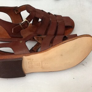 May include: Brown leather sandals with multiple straps and a buckle closure. The sandals have a flat sole and a small heel. The leather appears to be well-worn, giving the sandals a vintage look. The sandals are displayed on a white surface.