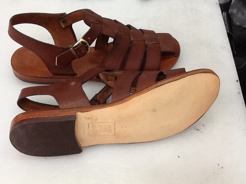 May include: Brown leather sandals with multiple straps and a buckle closure. The sandals have a flat sole and a small heel. The leather appears to be well-worn, giving the sandals a vintage look. The sandals are displayed on a white surface.