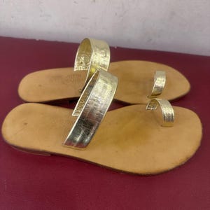 May include: A pair of flat sandals featuring a metallic gold finish on the straps and toe loops. The footbeds are tan and include a stamped logo that reads "MITIN SANDALS". The design includes a wide strap across the midfoot and a separate loop for the big toe.