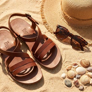 May include: Brown leather sandals with multiple straps, a straw hat, and tortoiseshell sunglasses rest on a sandy surface. The sandals are branded with "The STABAS" on the footbed. Seashells and small stones are scattered around the sandals.