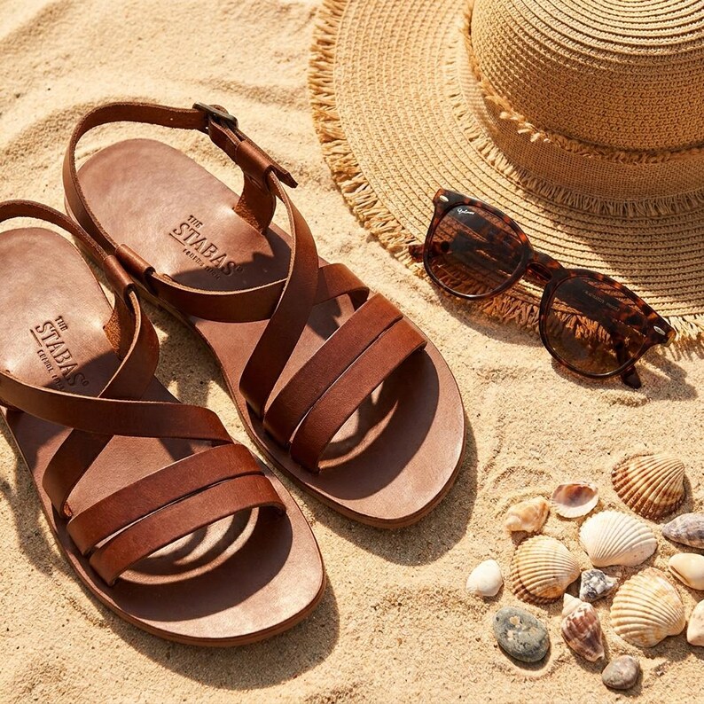 May include: Brown leather sandals with multiple straps, a straw hat, and tortoiseshell sunglasses rest on a sandy surface. The sandals are branded with "The STABAS" on the footbed. Seashells and small stones are scattered around the sandals.