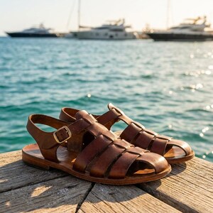May include: A pair of brown leather sandals with a woven design. The sandals have a buckle closure and are set on a wooden dock with the ocean and boats in the background.