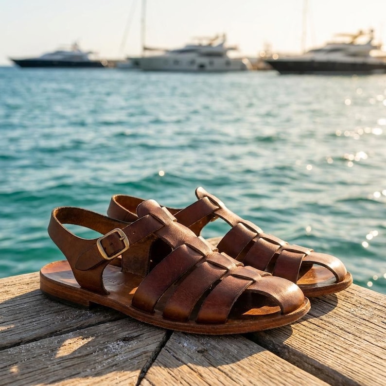 May include: A pair of brown leather sandals with a woven design. The sandals have a buckle closure and are set on a wooden dock with the ocean and boats in the background.