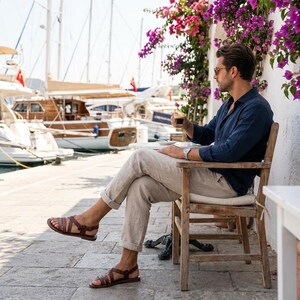 May include: A person wearing brown leather sandals with multiple straps, sitting on a wooden chair. The person is wearing a navy blue shirt and light-colored pants. The background includes boats, a harbor, and purple flowers.