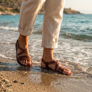 May include: Brown leather sandals with multiple straps. The sandals are worn on a sandy beach with the ocean in the background. The person is wearing beige pants.