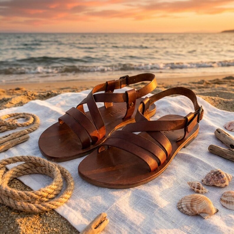 May include: A pair of brown leather sandals with multiple straps and buckles, resting on a white cloth on a sandy beach. The sandals are next to a coiled rope, driftwood, and seashells, with the ocean and a sunset in the background.