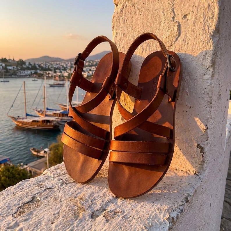 May include: A pair of brown leather sandals with multiple straps and buckle closures. The sandals are displayed on a white stone surface with a scenic background of boats on the water and a sunset.
