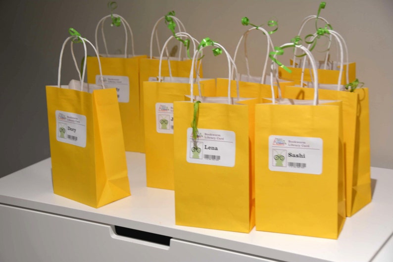 May include: Nine yellow paper gift bags with white handles and green ribbon. Each bag has a personalized label with a cartoon character wearing glasses and a name. The names on the labels are: Dory, Lena, Sashi.