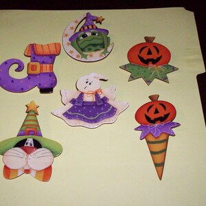 May include: Set of seven Halloween-themed wooden ornaments. The ornaments feature a witch's hat, a ghost, a pumpkin, a frog, a boot, and two pumpkins. The ornaments are painted in various colors, including green, orange, purple, and white.
