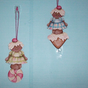May include: Two handmade wooden ornaments, each featuring a gingerbread girl doll. The top ornament is a stack of three pieces: a gingerbread girl doll, a white and pink frosting ice cream cone, and a brown waffle cone. The bottom ornament is a gingerbread girl doll standing on a pink and white candy swirl.