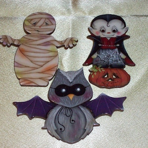 May include: Three Halloween-themed wooden ornaments: a mummy, a vampire, and a bat. The mummy is wrapped in white bandages with a sad expression. The vampire is wearing a red cape and has a mischievous grin. The bat has purple wings and a black mask.