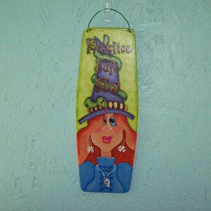 Puede incluir: Un letrero de madera con una bruja pintada que lleva un sombrero verde, morado y negro con las palabras "Toil & Trouble" en el letrero.