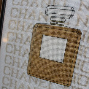 K&ouml;nnte beinhalten: Kreuzstich-Stickerei einer braunen Parf&uuml;mflasche mit wei&szlig;em Etikett und dem Wort "CHANEL" im Hintergrund.