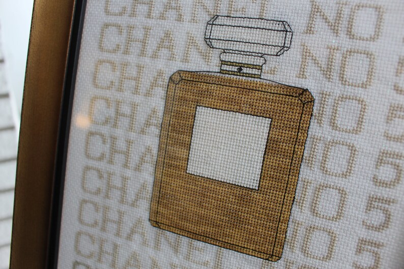 K&ouml;nnte beinhalten: Kreuzstich-Stickerei einer braunen Parf&uuml;mflasche mit wei&szlig;em Etikett und dem Wort "CHANEL" im Hintergrund.