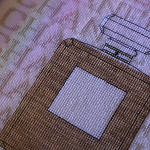 K&ouml;nnte beinhalten: Ein braunes und wei&szlig;es Kreuzstich-Design einer Parf&uuml;mflasche mit einer quadratischen Flasche und einem rechteckigen Verschluss.