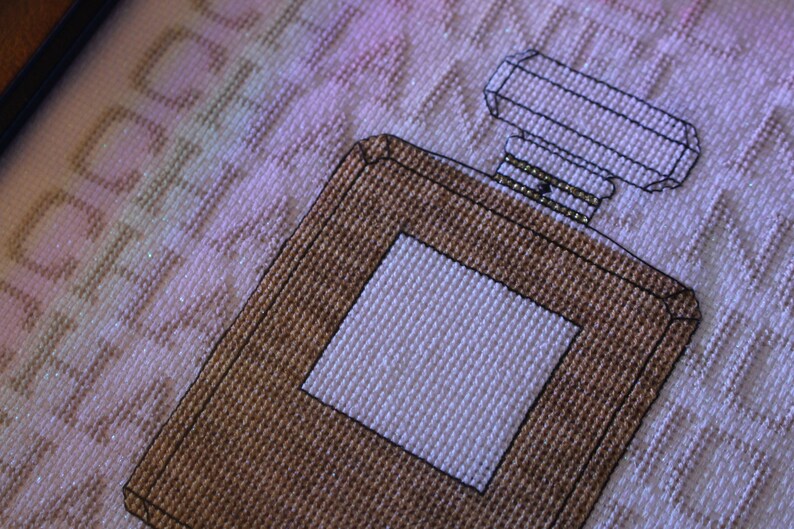 K&ouml;nnte beinhalten: Ein braunes und wei&szlig;es Kreuzstich-Design einer Parf&uuml;mflasche mit einer quadratischen Flasche und einem rechteckigen Verschluss.