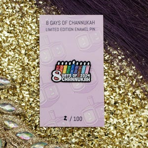 May include: A pink enamel pin with a rainbow design and the text "8 Days of 2024 Chanukah". The pin is a limited edition and is numbered 2/100.
