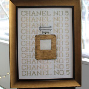K&ouml;nnte beinhalten: Ein gerahmtes Kreuzstich-Design mit einem goldenen Parf&uuml;mflakon mit dem Text "CHANEL NO 5", der mehrmals wiederholt wird.