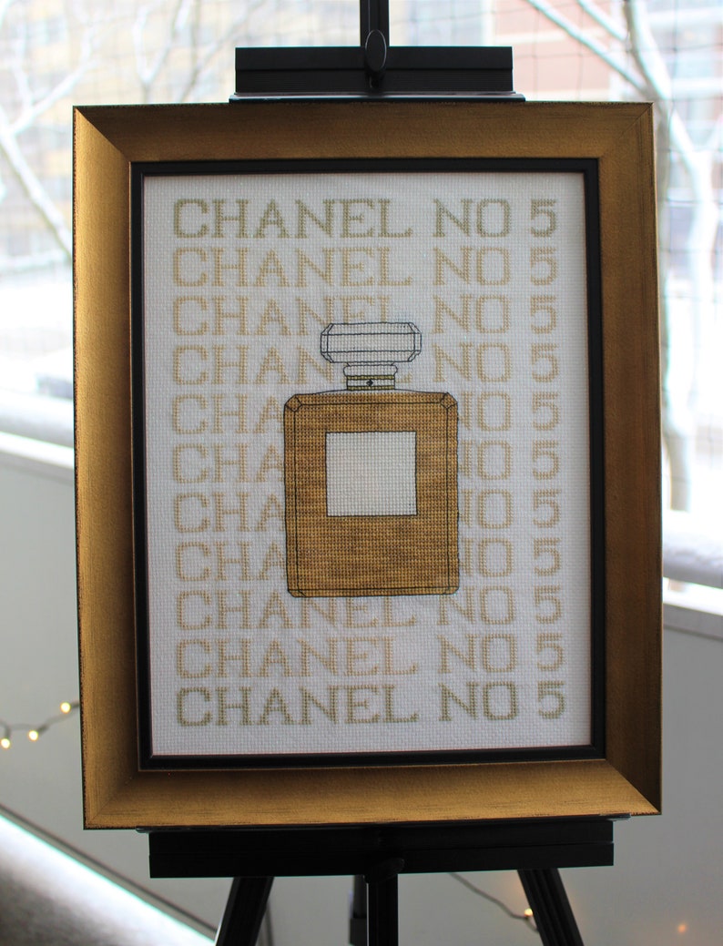 K&ouml;nnte beinhalten: Ein gerahmtes Kreuzstich-Design mit einem goldenen Parf&uuml;mflakon mit dem Text "CHANEL NO 5", der mehrmals wiederholt wird.
